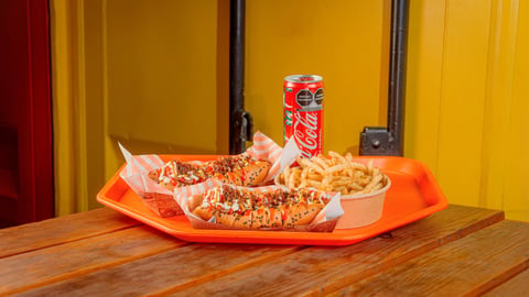 Orange tray with two sandwiches wrapped in paper, french fries, and a cola can on a wooden table against yellow wall
