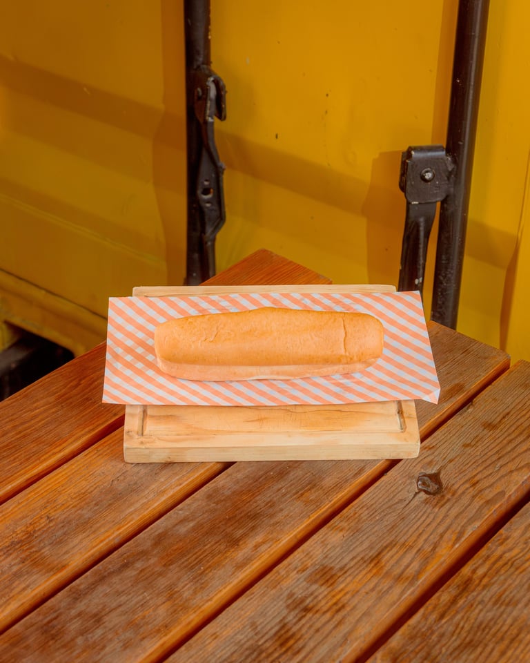 A fresh baguette on striped orange and white paper sitting on a wooden board against a bright yellow background