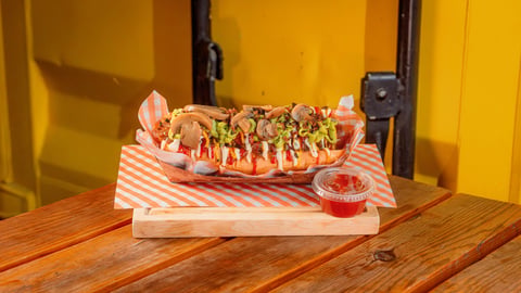 Loaded hot dog with toppings on striped paper, served on wooden table with yellow background and small condiment cup