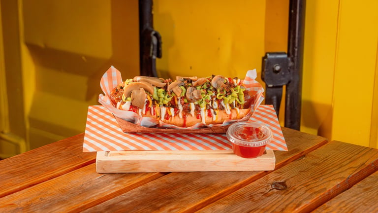 Loaded hot dog with toppings on striped paper, served on wooden table with yellow background and small condiment cup