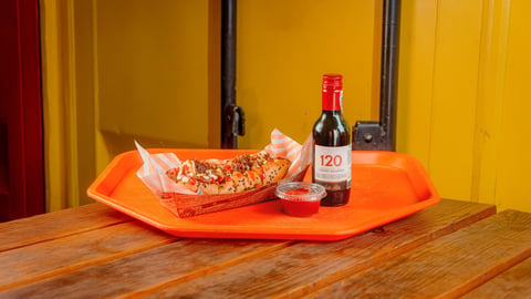 Hot dog in foil wrapper on orange tray with wine bottle and glass on wooden table against yellow wall